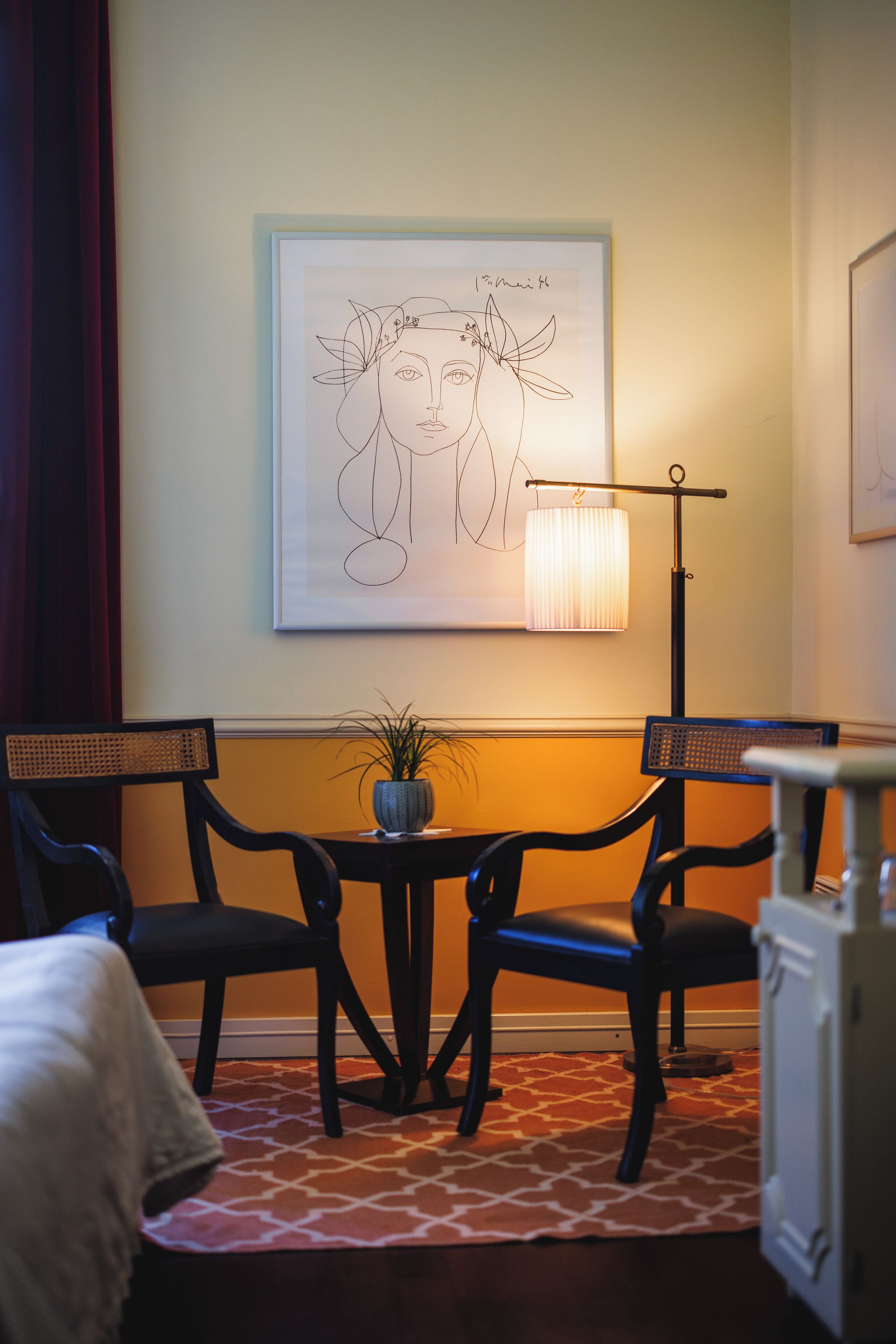 Habitación de hotel con una cama, dos sillas, una mesita con una planta, una obra de arte enmarcada en la pared y una lámpara de pie.