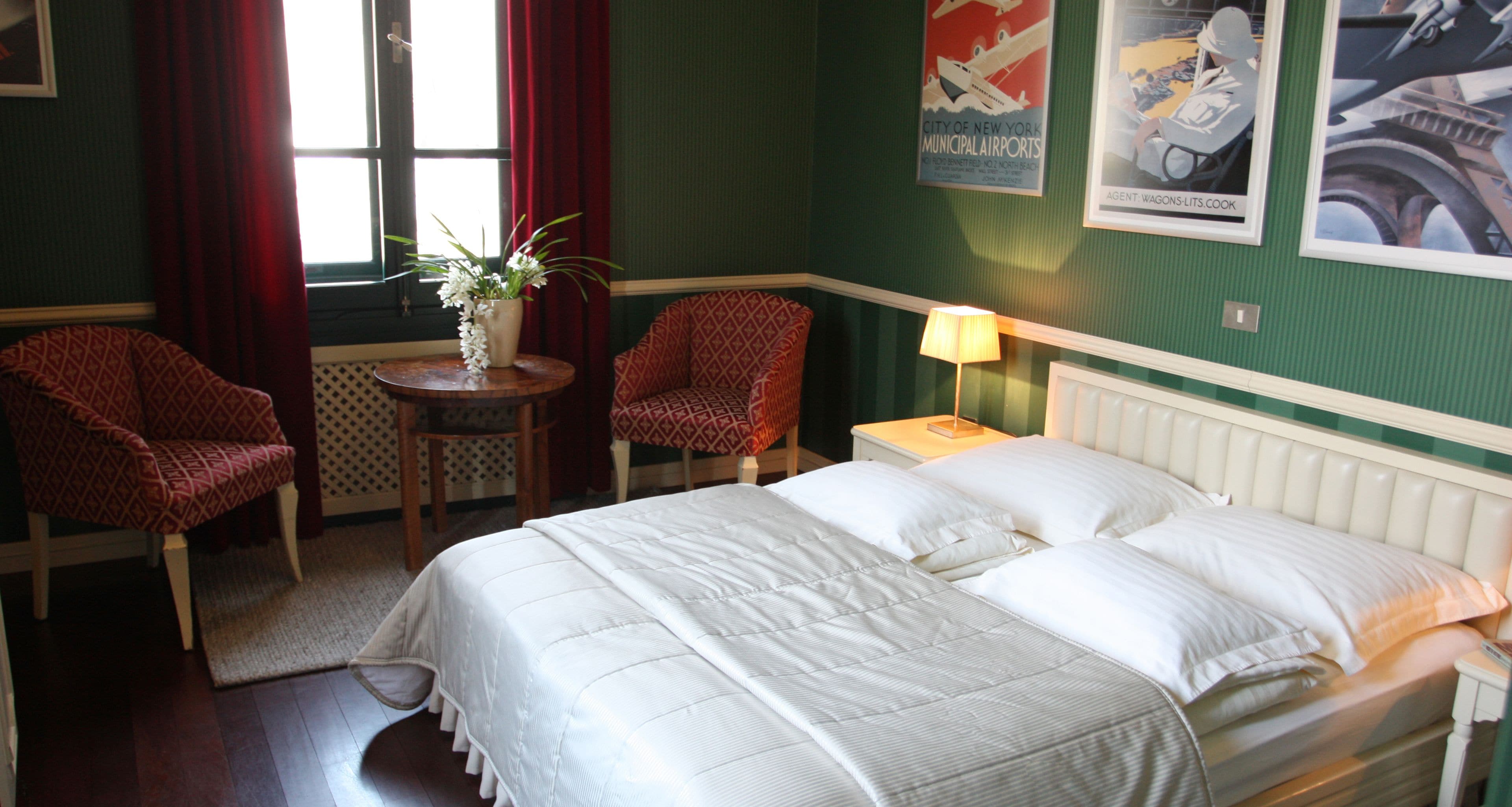 A hotel room with a large bed, two armchairs, a small table with a vase of flowers, and a window with red curtains.