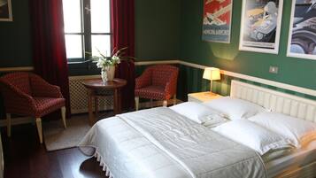 A hotel room with a large bed, two armchairs, a small table with a vase of flowers, and a window with red curtains.