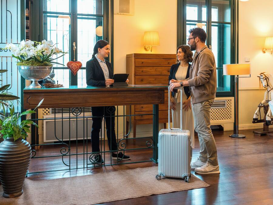 A reception desk with a receptionist, a guest with a suitcase, and a man conversing.