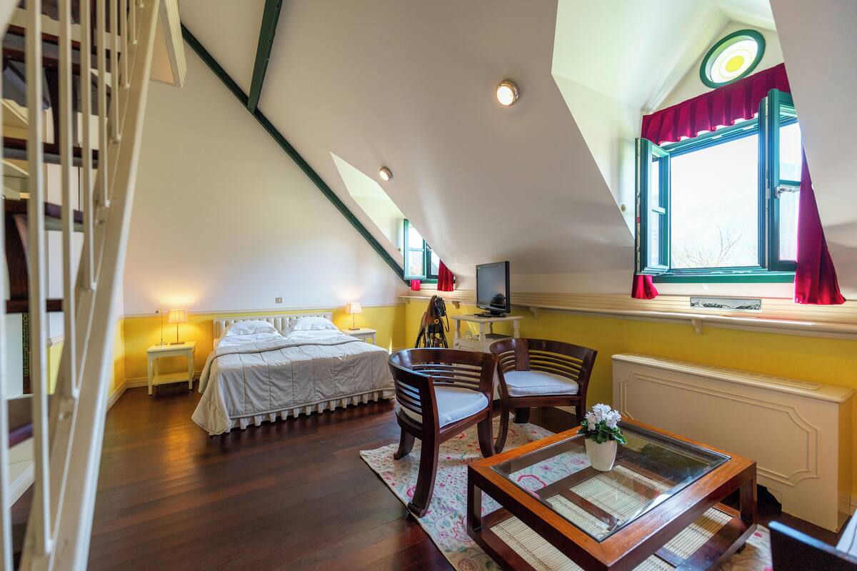 A bedroom with a bed, two chairs, a table, a TV, and a window with curtains.