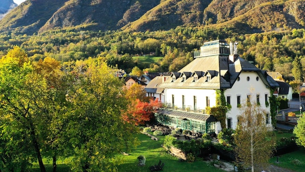 Un edificio de estilo chalé tradicional con techo inclinado, rodeado de exuberante vegetación y montañas al fondo.