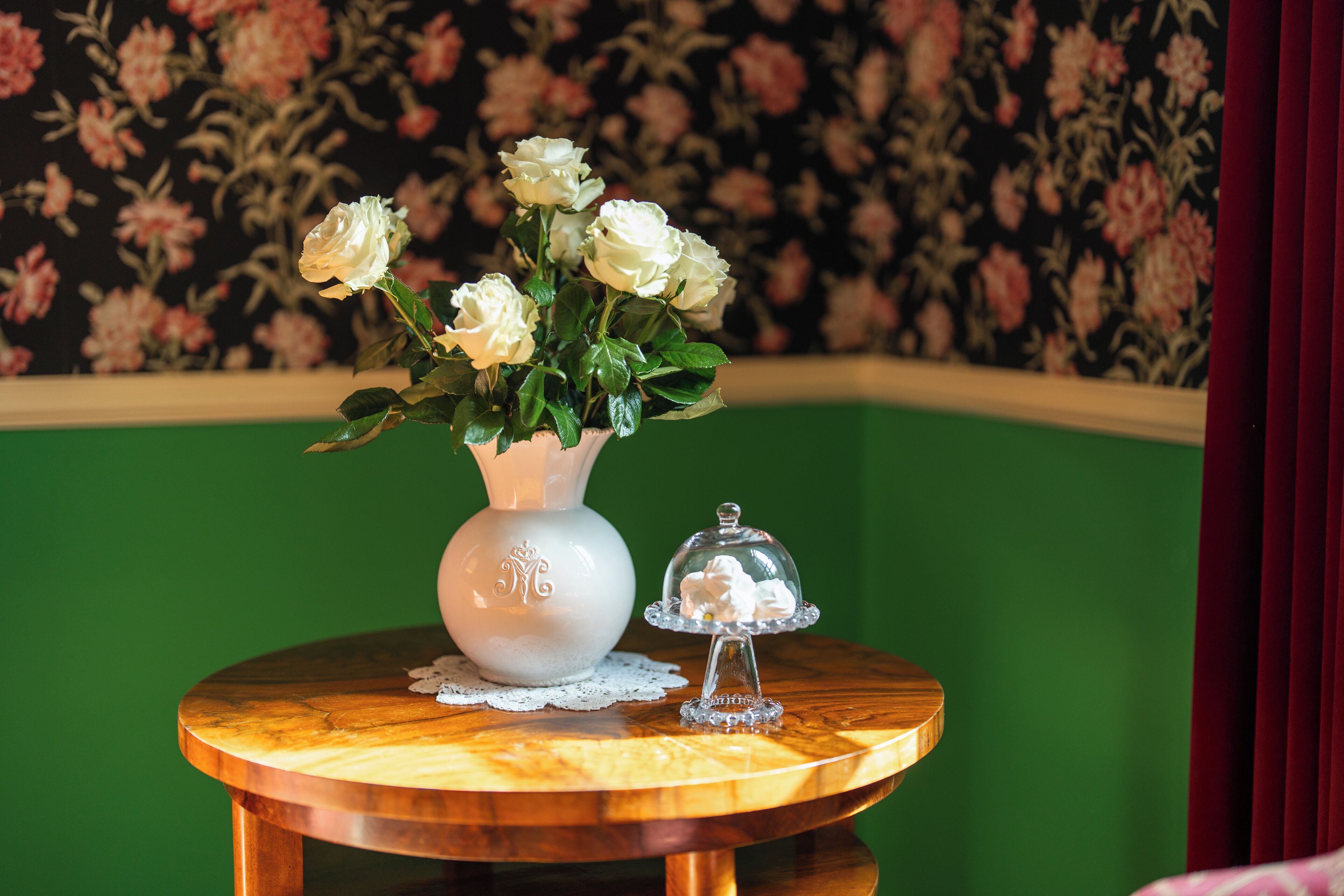 A round wooden table with a white vase containing white flowers, a glass dome with cream-colored items, and a floral wallpaper background.