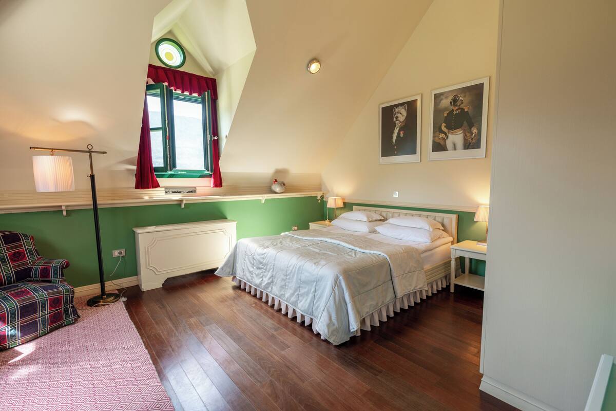 A bedroom with a bed, a nightstand, a lamp, a chair, a window with curtains, and two framed pictures on the wall.