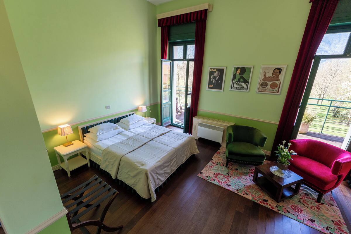 A bedroom with a bed, a nightstand, a chair, a table with a plant, and a window with curtains.