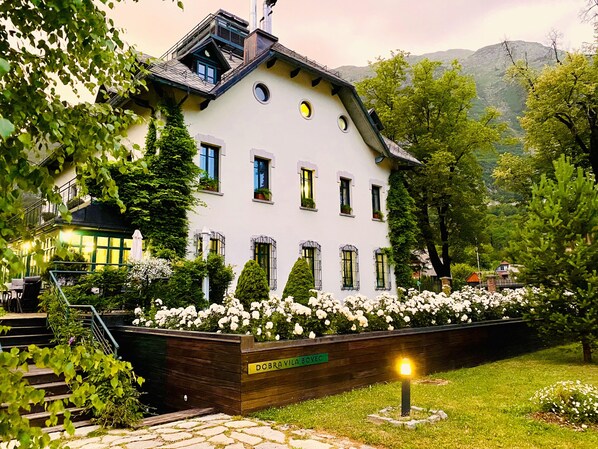A white building with multiple windows, surrounded by greenery and a stone pathway. - Hotel Dobra Vila (Bovec)