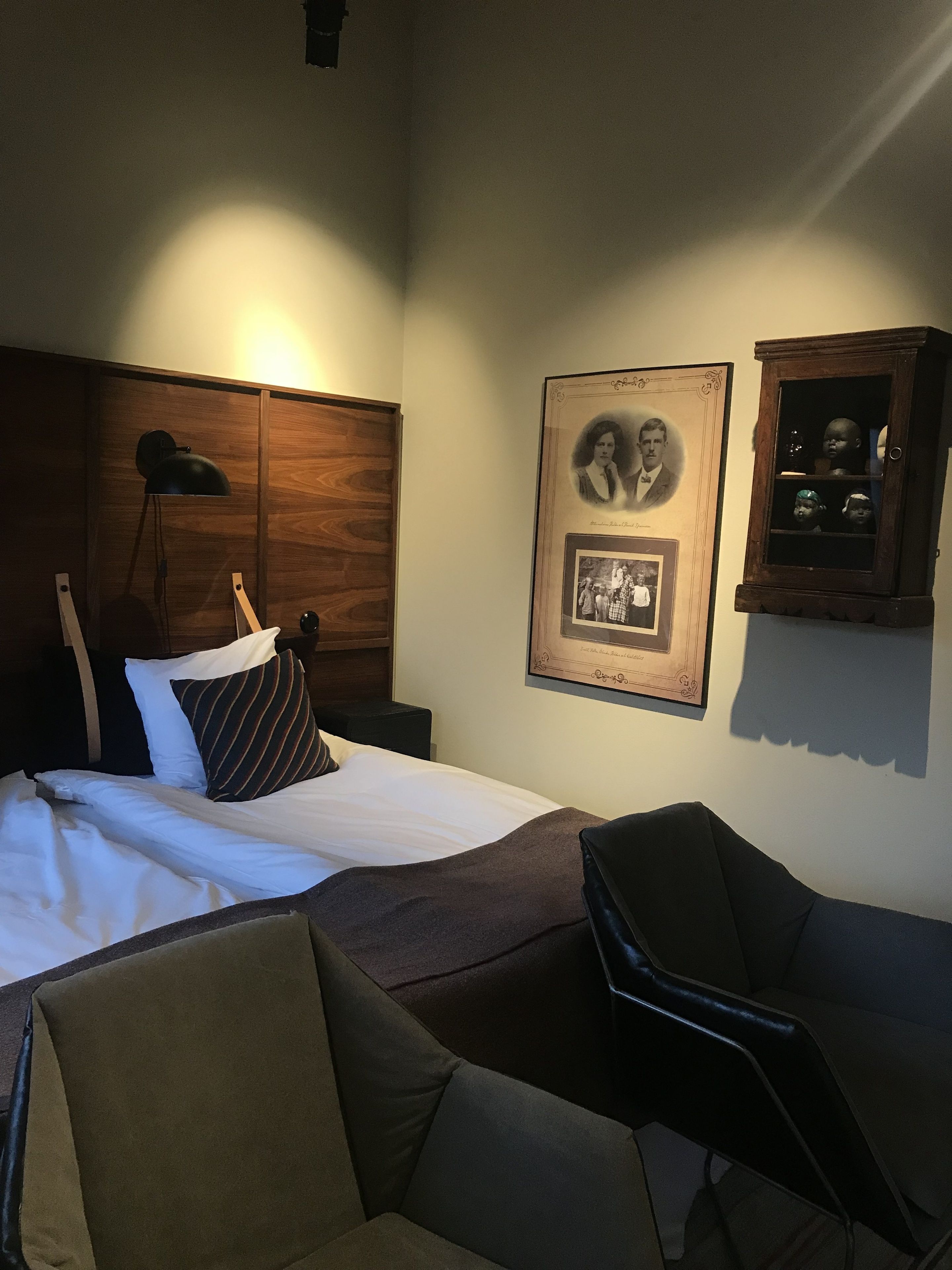 A hotel room with a bed, a desk, a chair, a lamp, and framed pictures on the wall.
