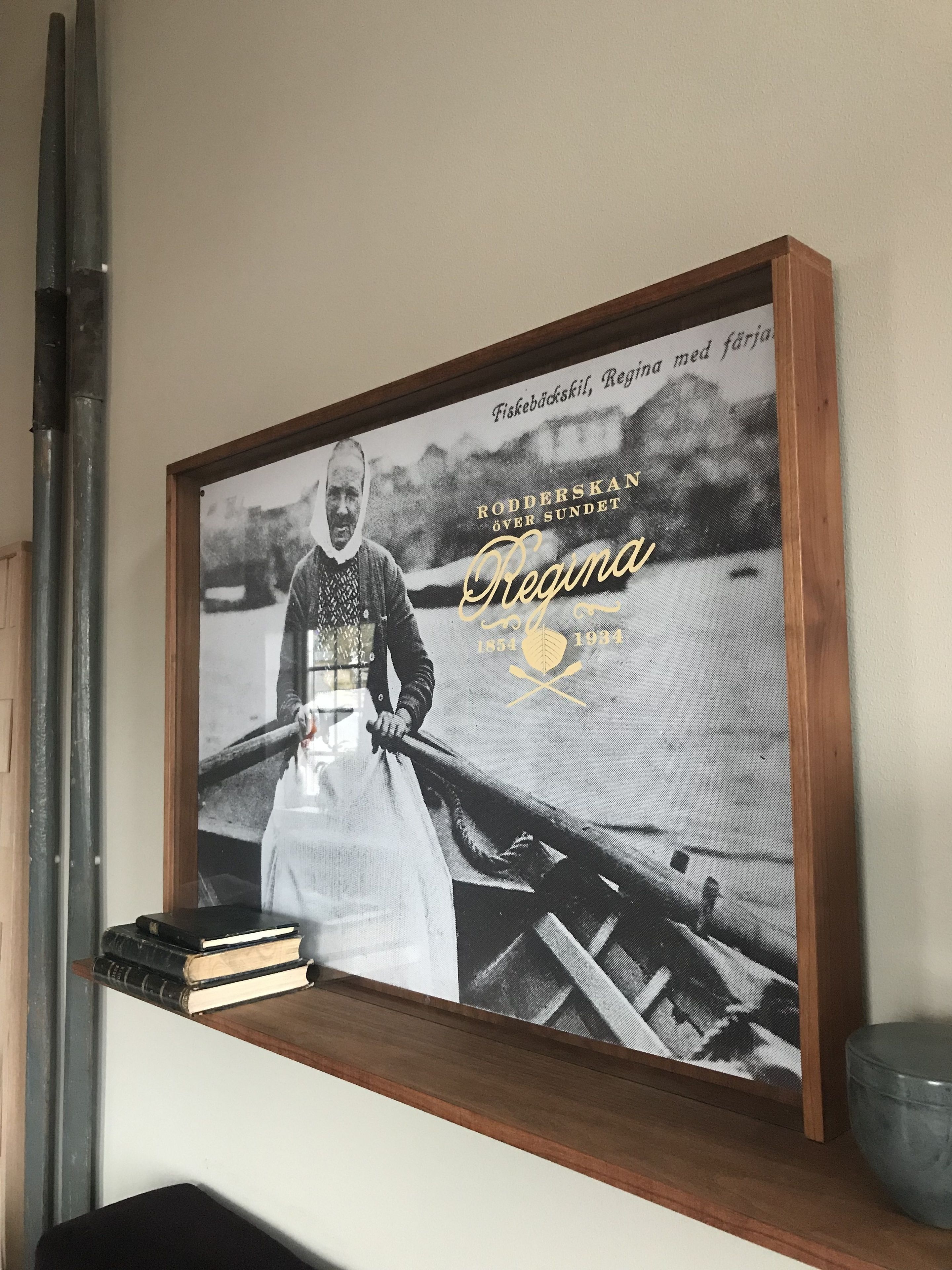 Fotografía histórica enmarcada con texto y un logotipo, ubicada en una estantería con libros y un pequeño recipiente.