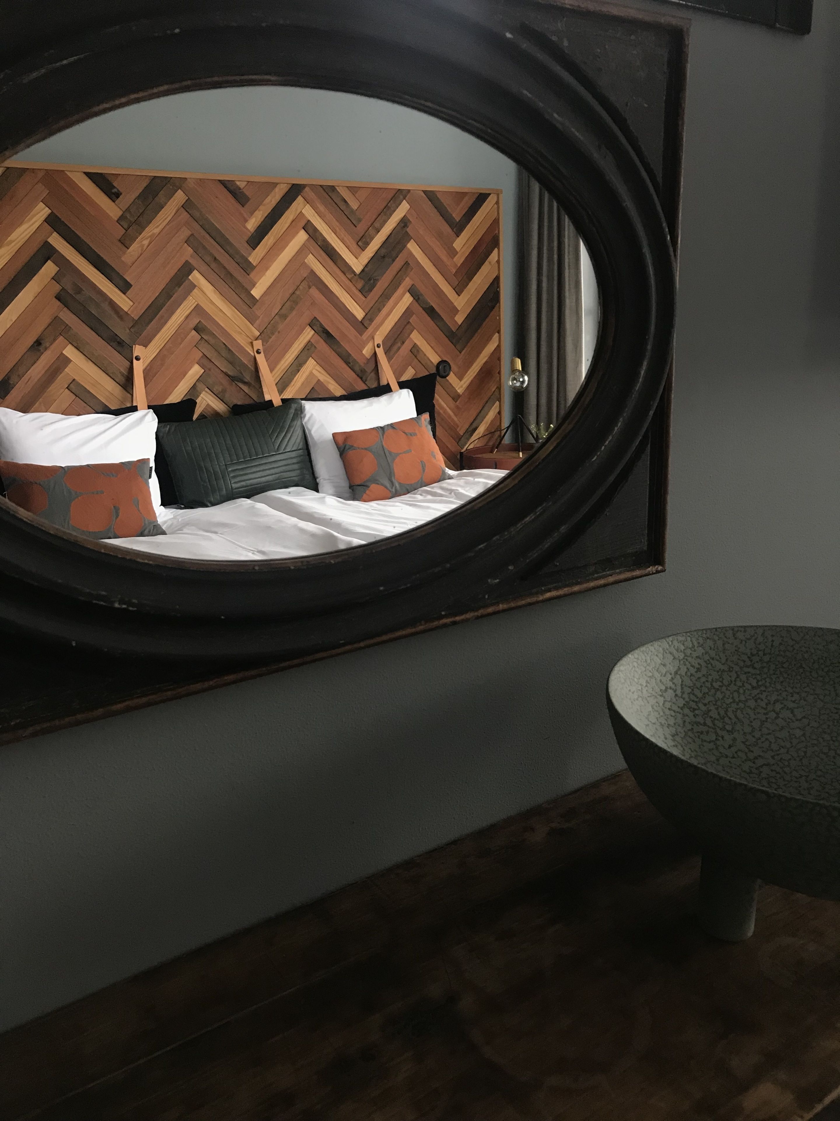 A bedroom with a circular mirror reflecting a bed with white bedding and orange patterned pillows. The room features a herringbone wood wall pattern and a modern, dark-toned sink.