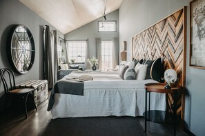 Een slaapkamer met een groot bed, een houten hoofdbord, een ronde spiegel, een dressoir en een raam met gordijnen.