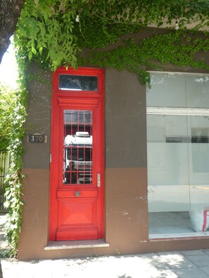 A red door with a window, a number plate, and a building with a glass window. - Departamento-casa muy amplio y céntrico. (Luján)