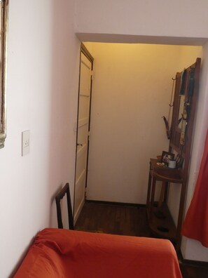 A small, dimly lit room with a bed, a chair, a desk with a mirror, and a door leading to another room. - Departamento-casa muy amplio y céntrico. (Luján)