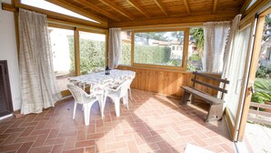 Une terrasse couverte avec une table et des chaises, des bancs en bois et de grandes fenêtres.
