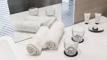 Un baño con toallas blancas, dos vasos de vidrio y dos botellas de artículos de tocador sobre una encimera blanca.