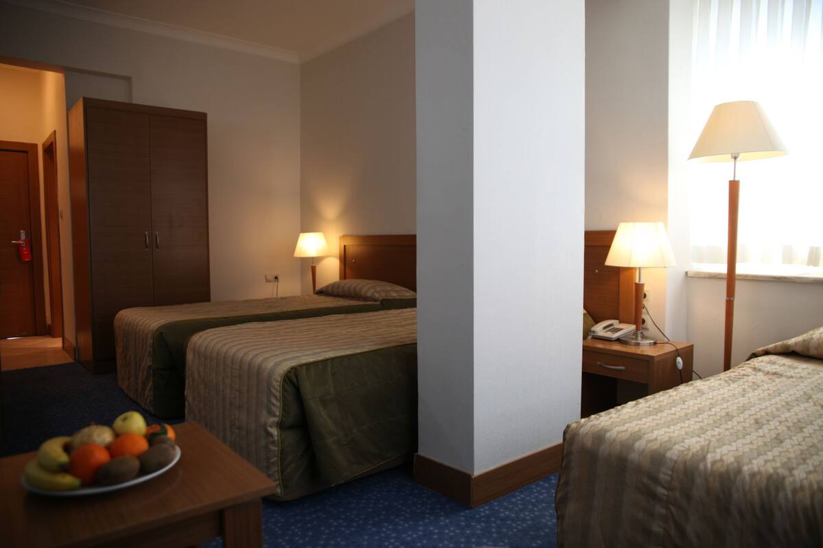 A hotel room with two single beds, a wooden wardrobe, a bedside table with a lamp, and a fruit bowl on the nightstand.