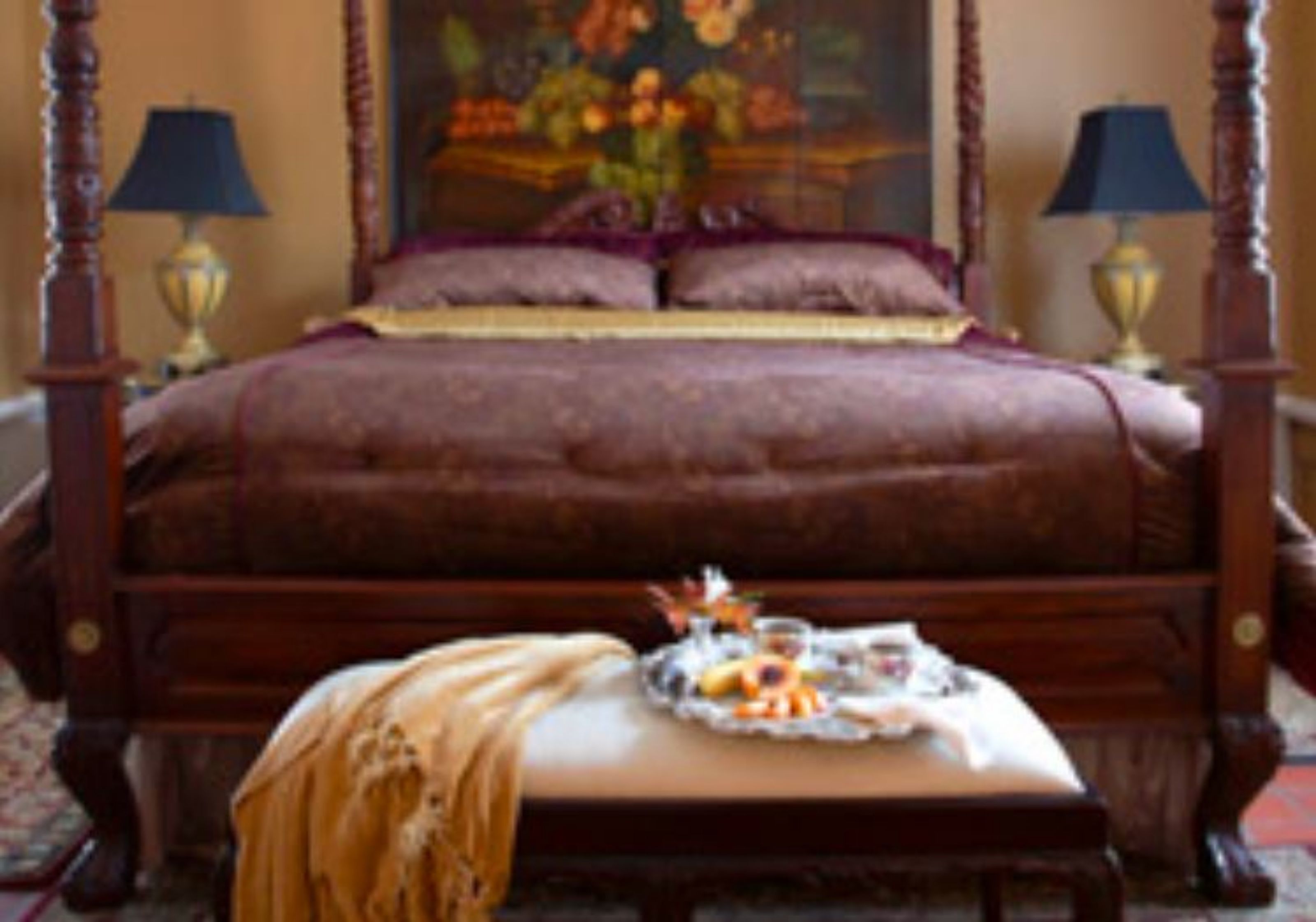 A four-poster bed with a floral painting above it, a bedside table with a lamp, and a tray with fruits and a teapot on the bed.