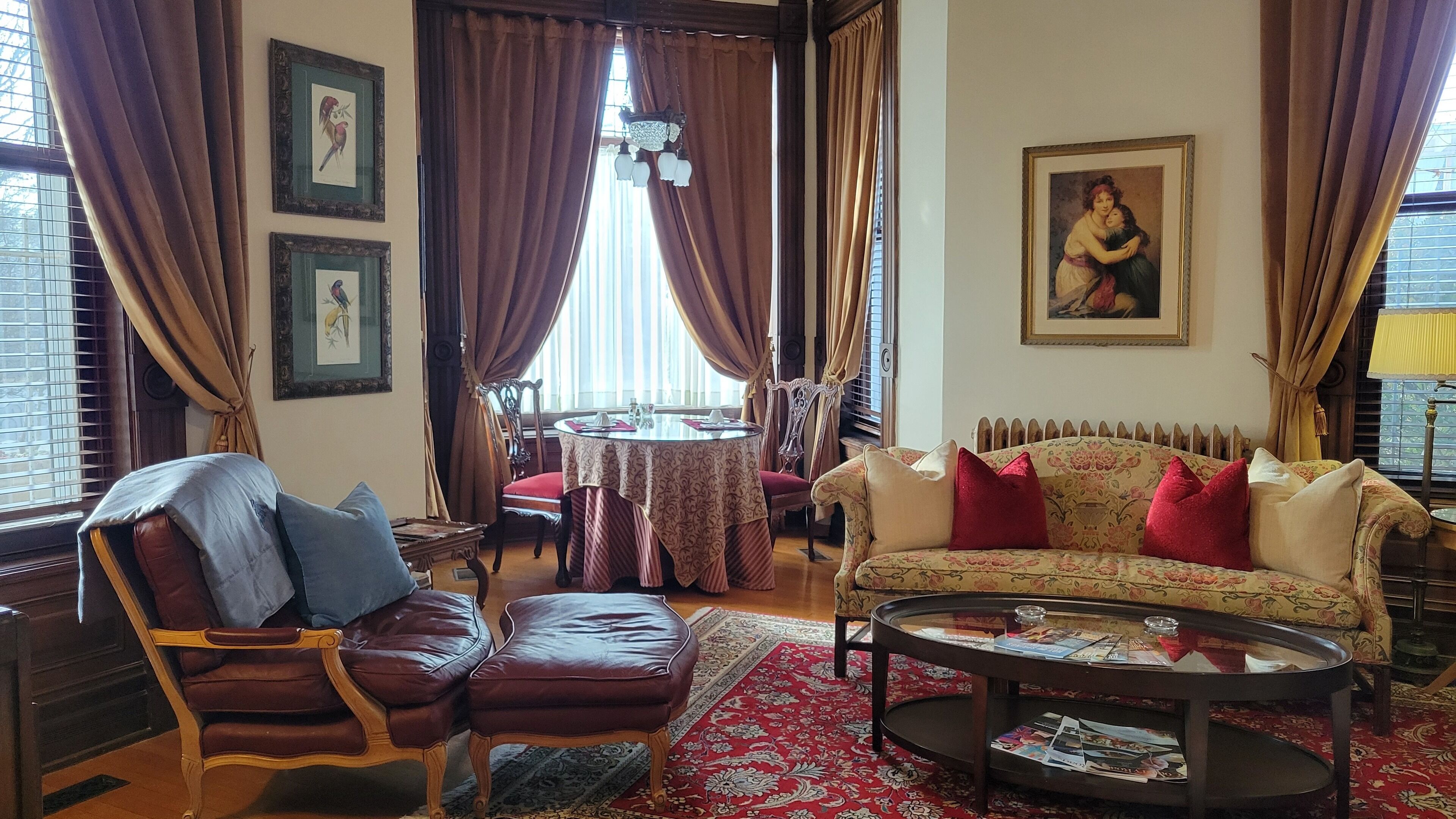 Un salón tradicional con un sillón de cuero, un sofá con cojines rojos, una mesa de centro redonda y un comedor con mesa y sillas.