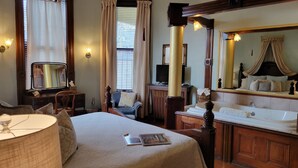 A hotel room with a bed, a desk, a television, a large mirror, a vanity with a bathtub, and a window with curtains. - 1890 Williams House Inn (Hot Springs)