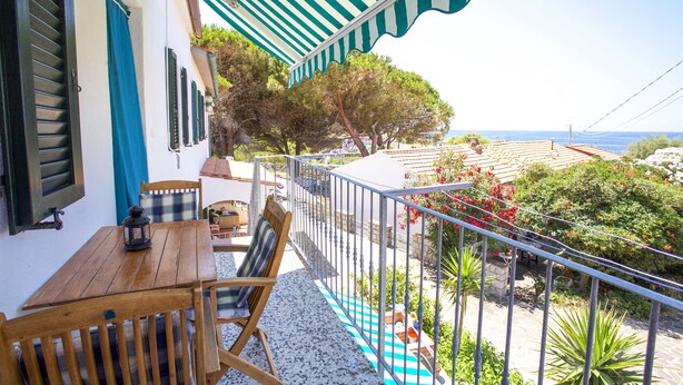A balcony with a wooden table and chairs, a striped awning, and a view of the sea and greenery. - Appartamenti Il Marino (Marciana)