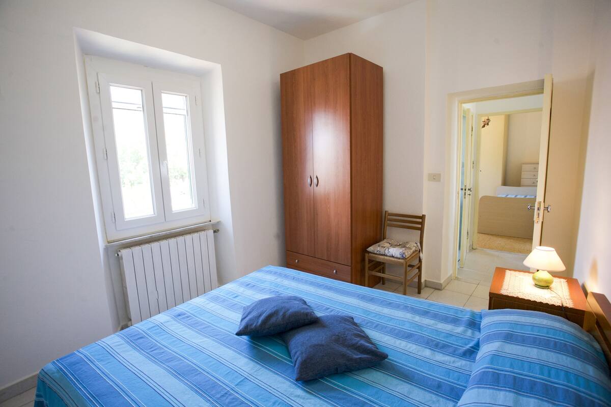 Quarto com cama, guarda-roupa de madeira, uma cadeira, mesa de cabeceira com abajur e janela.