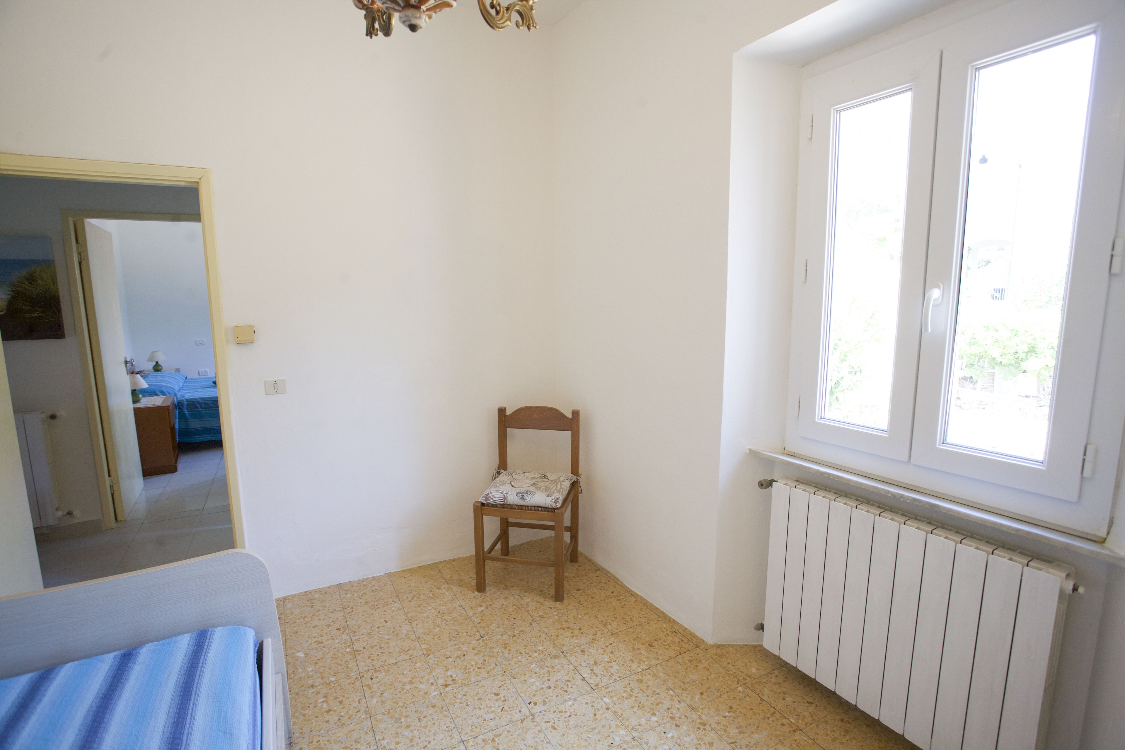 A room with a wooden chair, a bed, a radiator, and a window with a view of greenery.