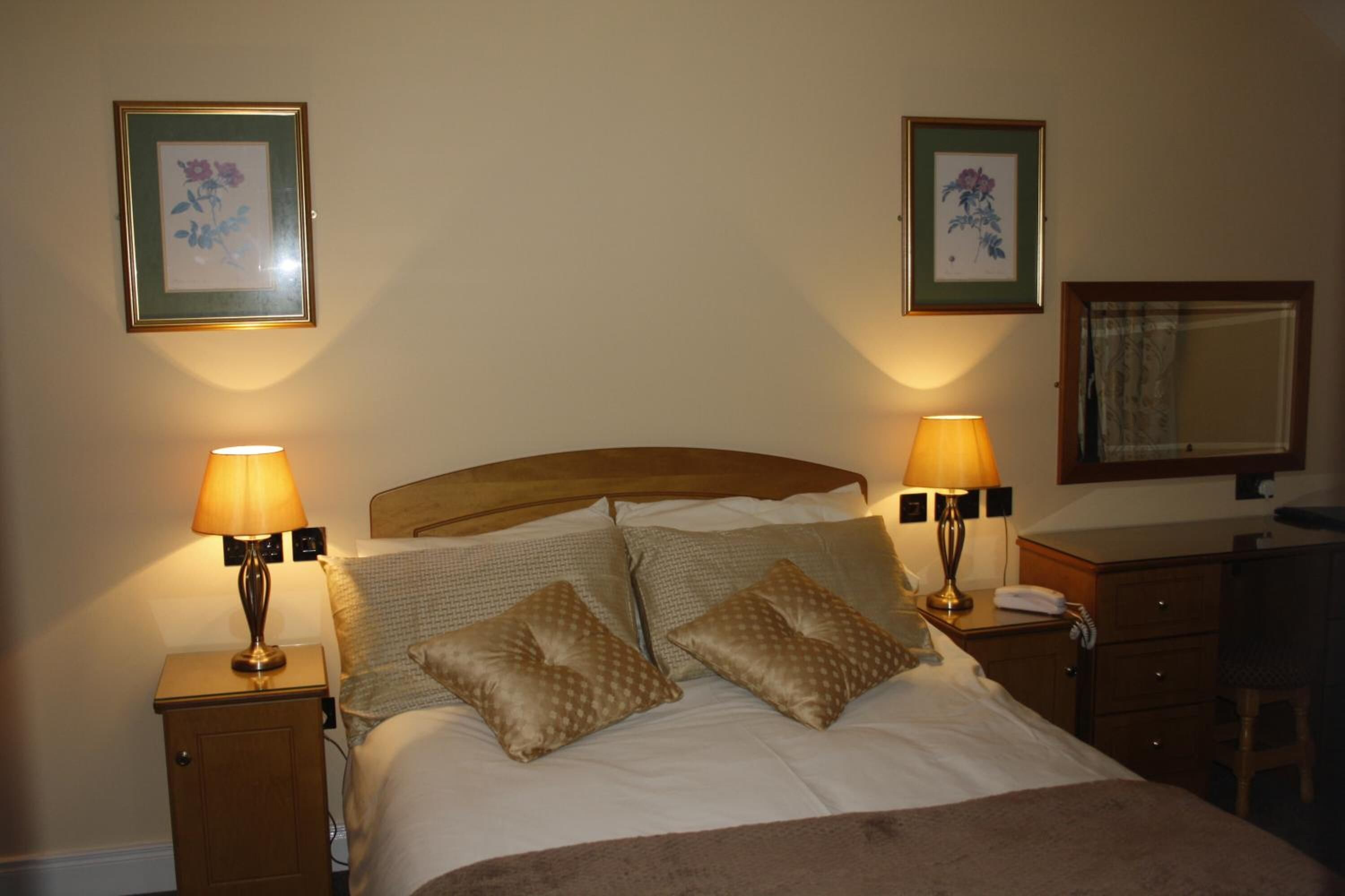 A neatly made bed with two pillows, two bedside lamps, a wooden nightstand, a wooden dresser, and two framed floral pictures on the wall.