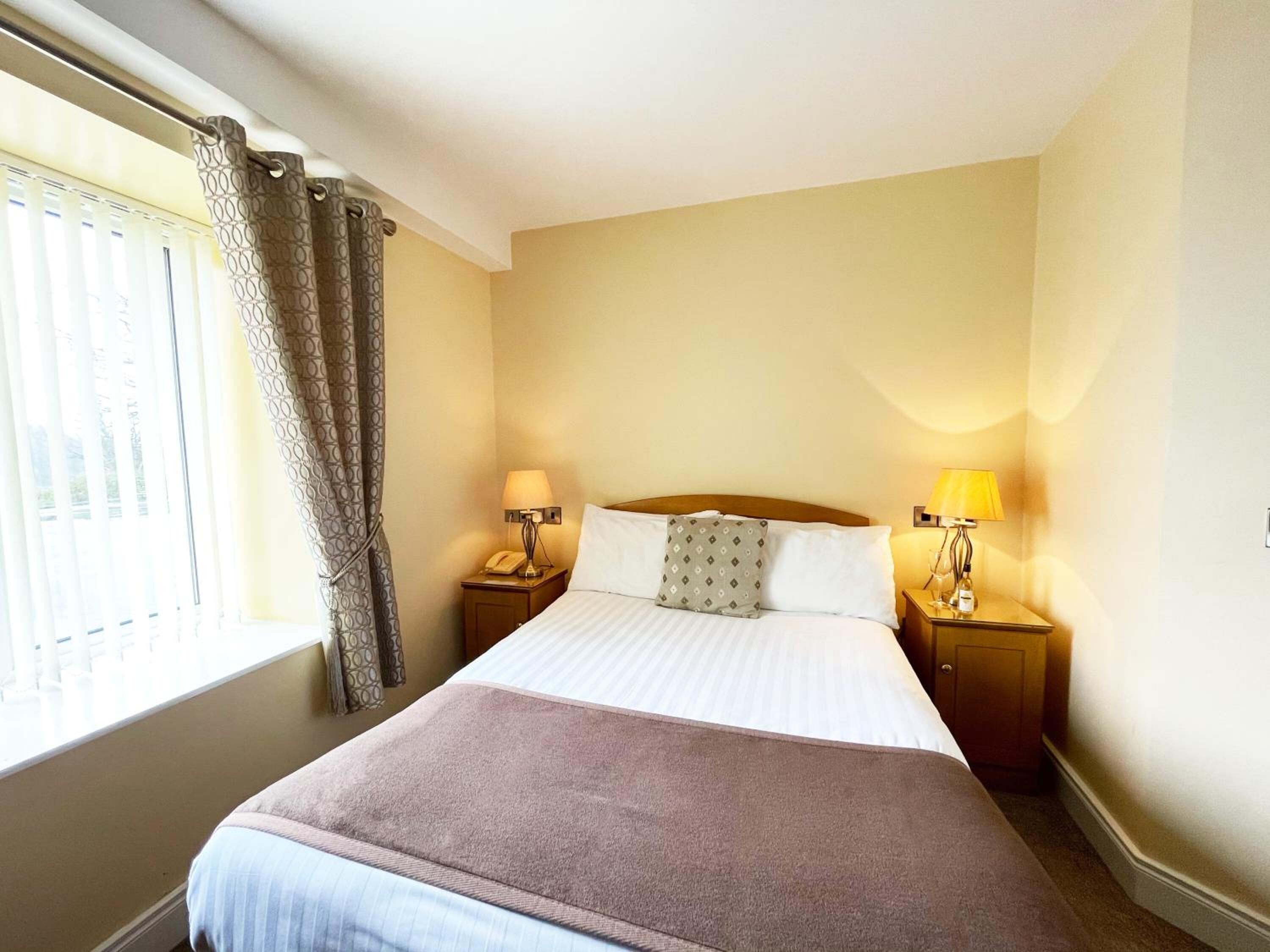 Ein Hotelzimmer mit einem Bett, zwei Nachttischen, einer Lampe, einem Telefon und einem Fenster mit Vorhängen.