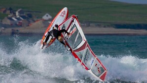 A windsurfing athlete performing a trick on a board with a red and white design, in a body of water with waves, and a distant shoreline with buildings.