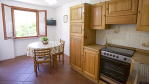 A kitchen with wooden cabinets, a stove, a dining table with chairs, and a window with curtains. - Appartamento Il Marino II (Marciana)