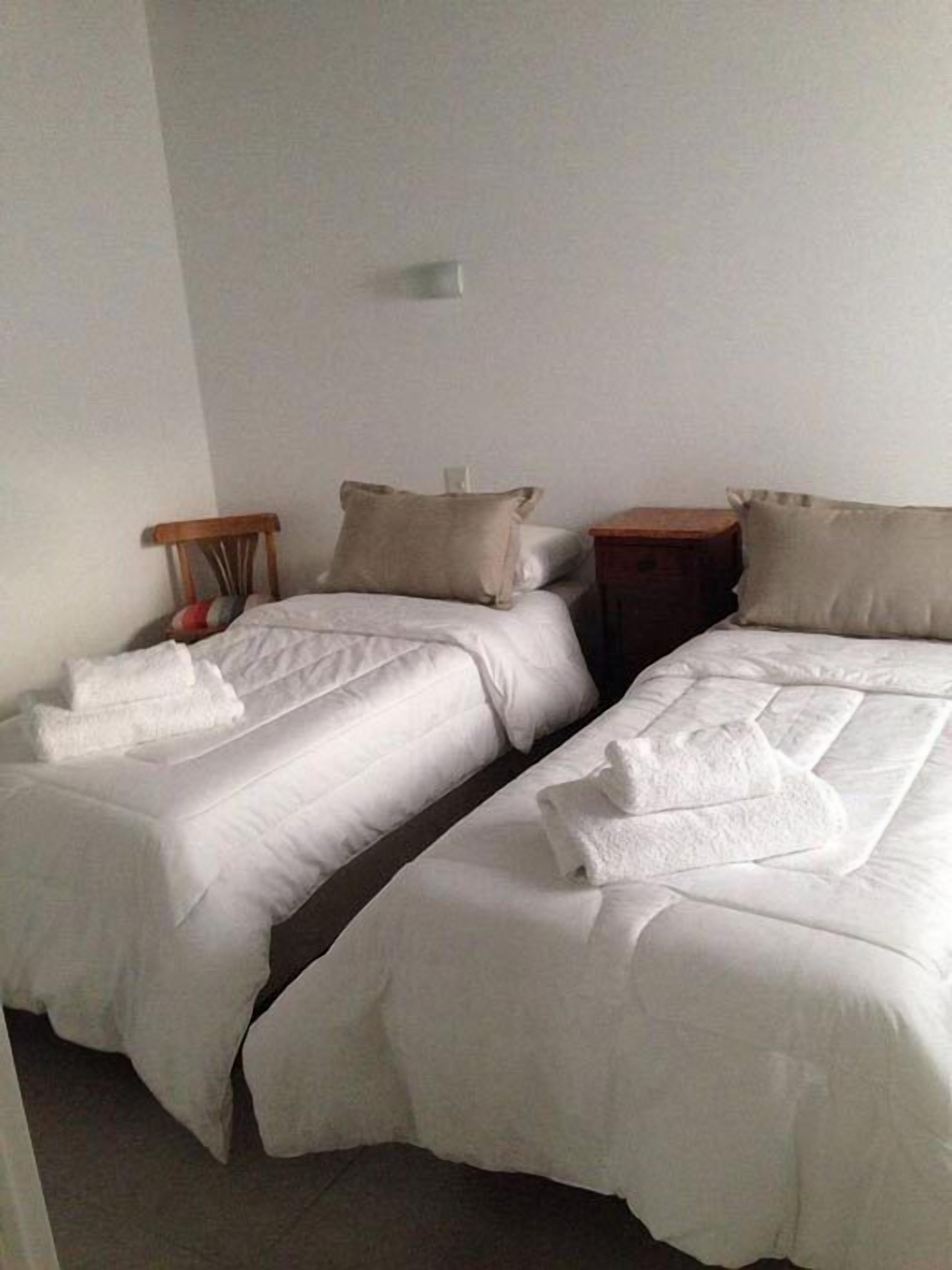 Dos camas individuales con ropa blanca, una mesita de noche de madera y una pequeña mesa con una lámpara.