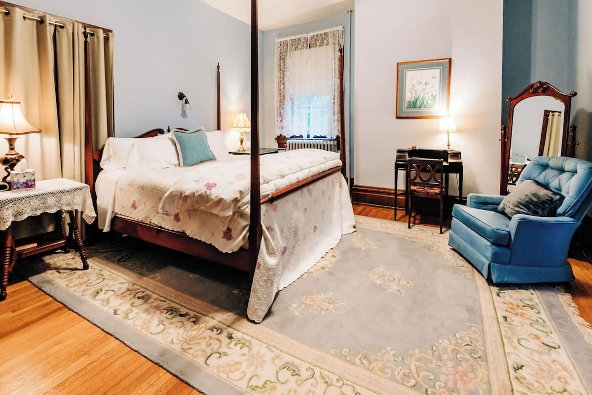 A bedroom with a four-poster bed, a blue armchair, a wooden desk, and a large mirror.