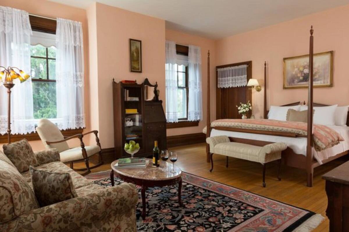 A traditional bedroom with a four-poster bed, a sitting area with a sofa and armchairs, a wooden side table, and a bookshelf.