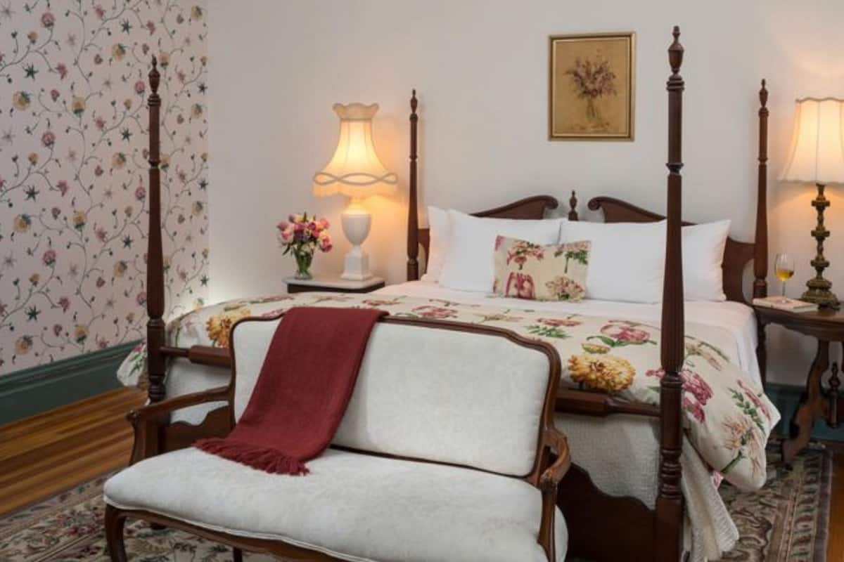A four-poster bed with floral bedding, a red throw blanket, and a wooden chair in a room with floral wallpaper and two bedside tables with lamps.