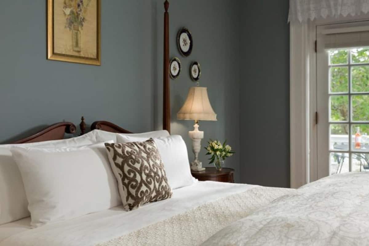 A neatly made bed with white linens and decorative pillows, a bedside table with a lamp and a vase of flowers, and a window with a view of greenery.