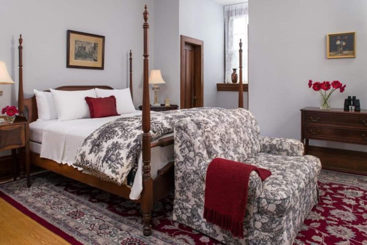 A four-poster bed with a floral bedspread, a wooden nightstand with a lamp, a chair with a red throw, and a dresser with a vase of flowers.