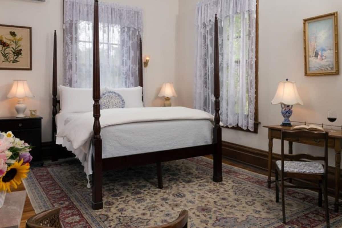 A four-poster bed with white linens, a floral-patterned rug, and a wooden nightstand with a lamp.