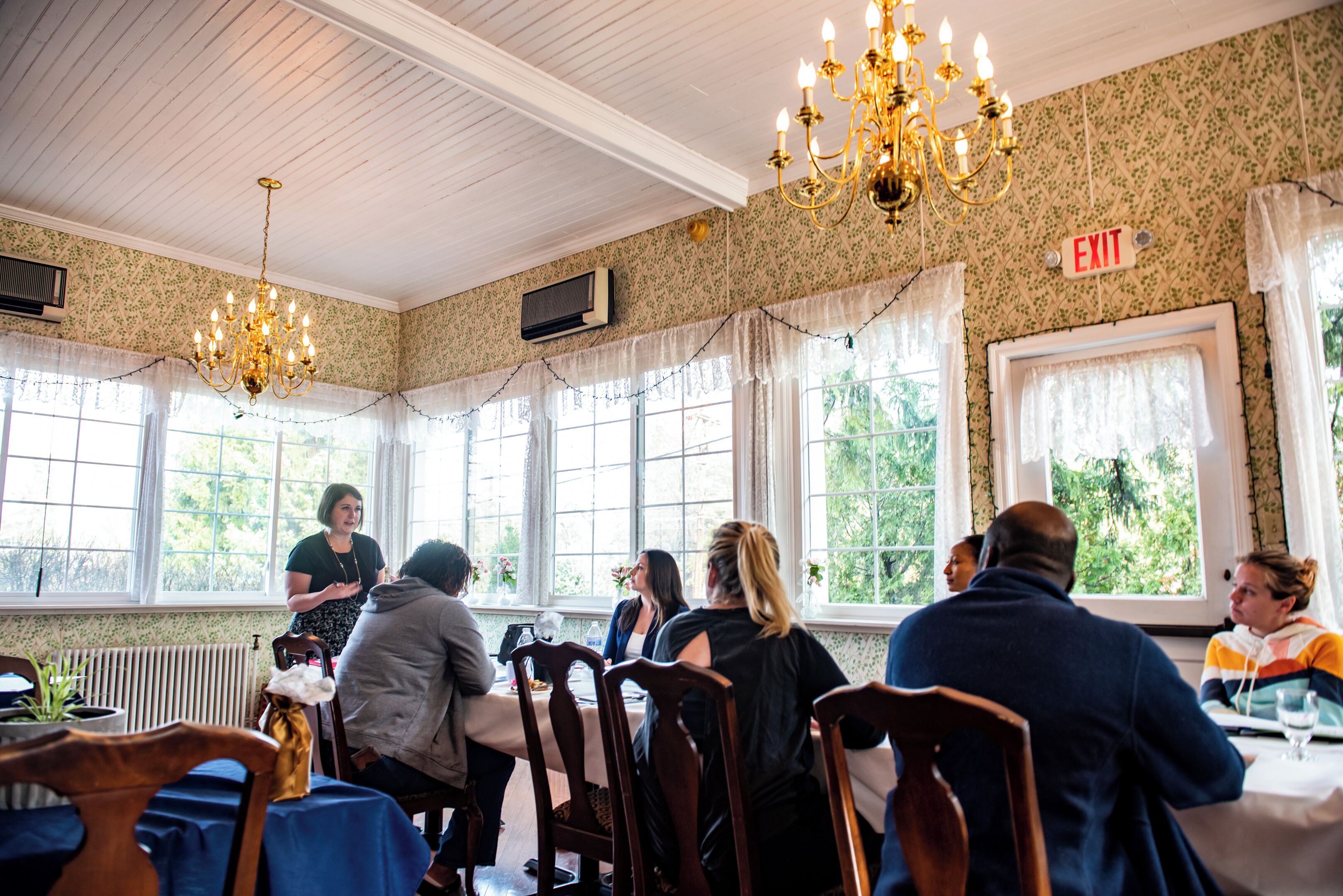 A dining area with wooden tables and chairs, chandeliers, and large windows with curtains.