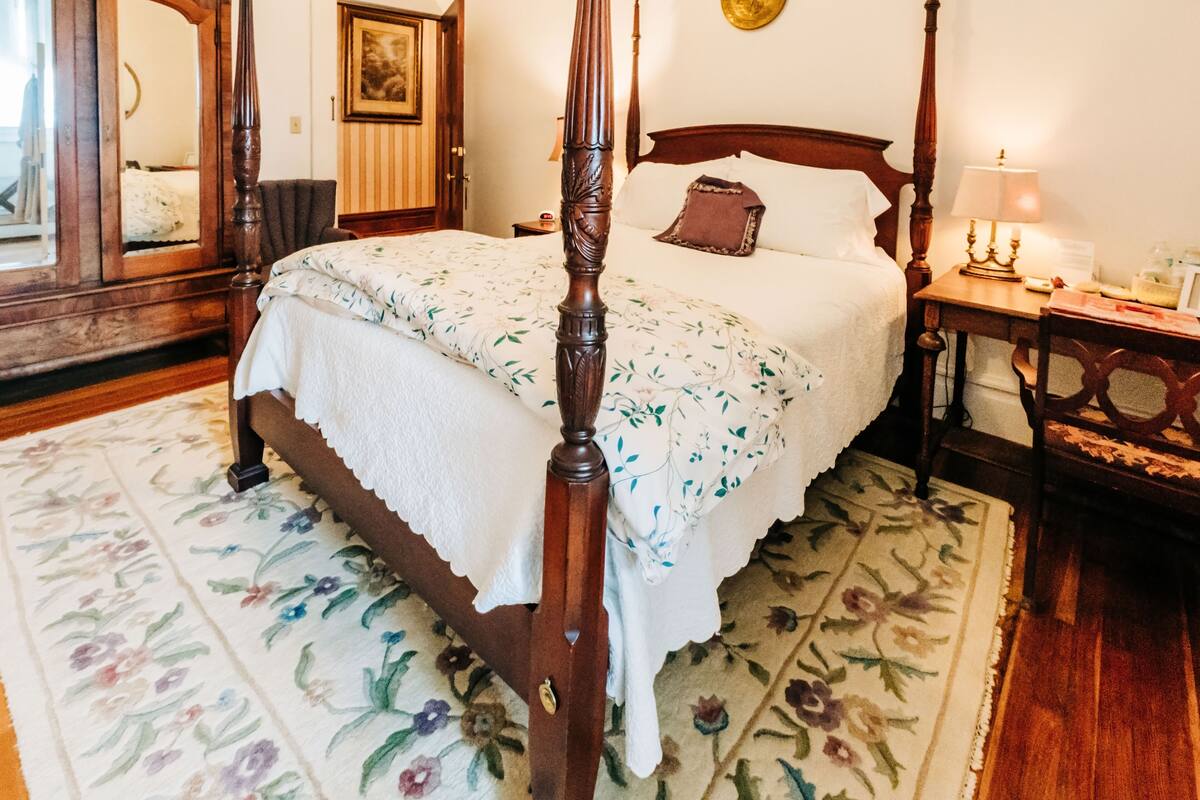 A four-poster bed with a floral bedspread, a wooden nightstand with a lamp, and a mirror on the wall.