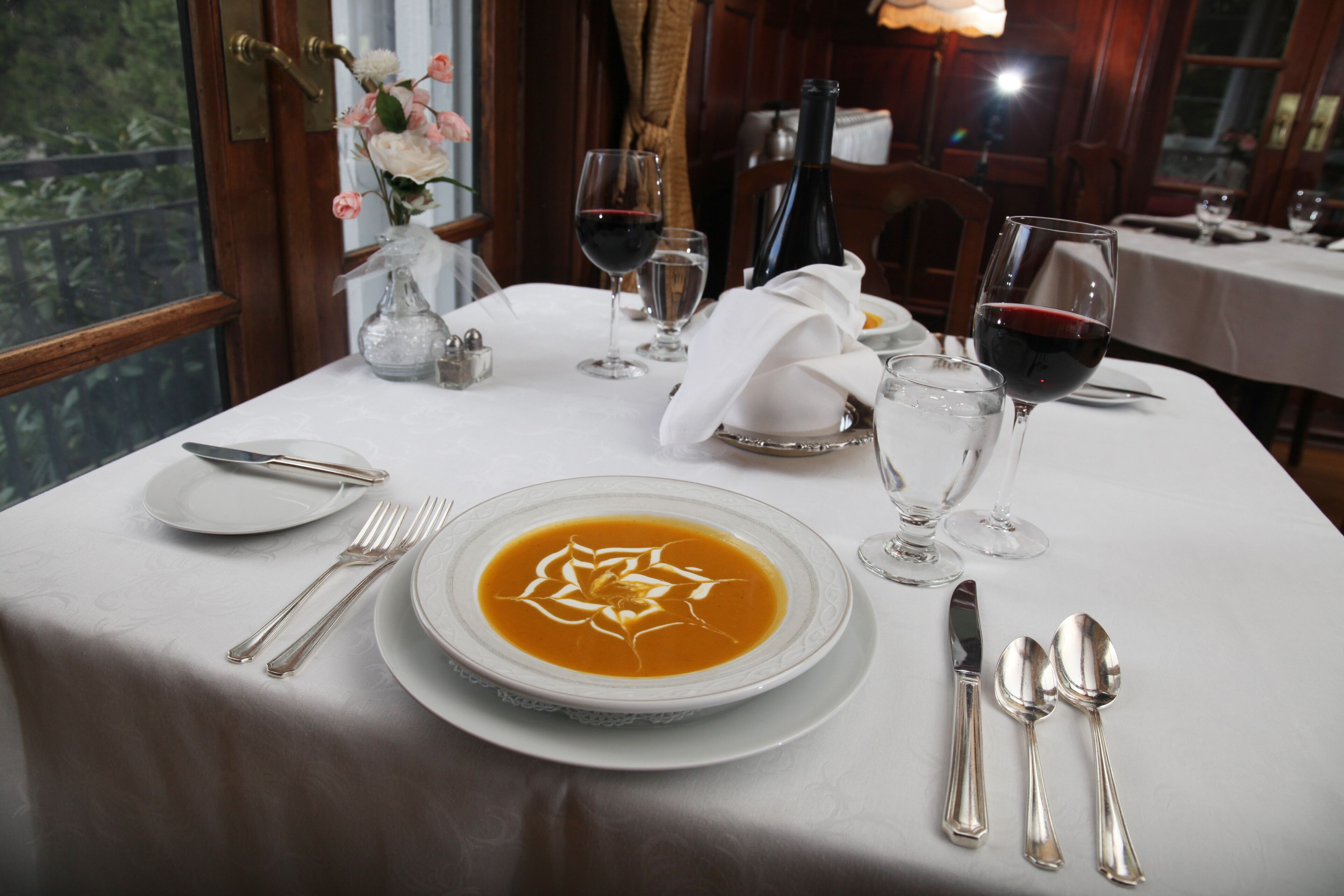 A dining table set with white tablecloth, silverware, a bowl of soup, wine glasses, and a vase with flowers.
