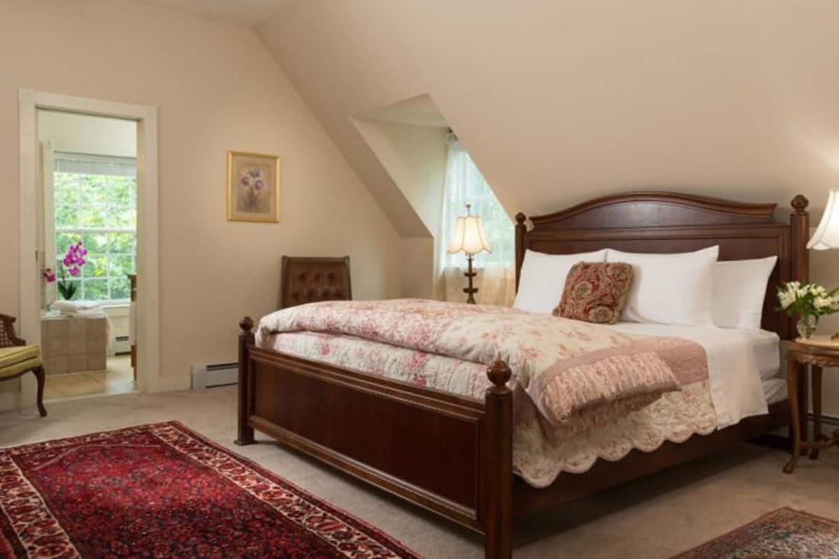 A bedroom with a wooden bed, two bedside lamps, a red and beige rug, and a window with a view of greenery.