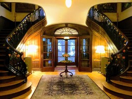 Una imponente escalera con barandillas ornamentadas y un candelabro central, que conduce a una puerta de madera con paneles de vidrio.