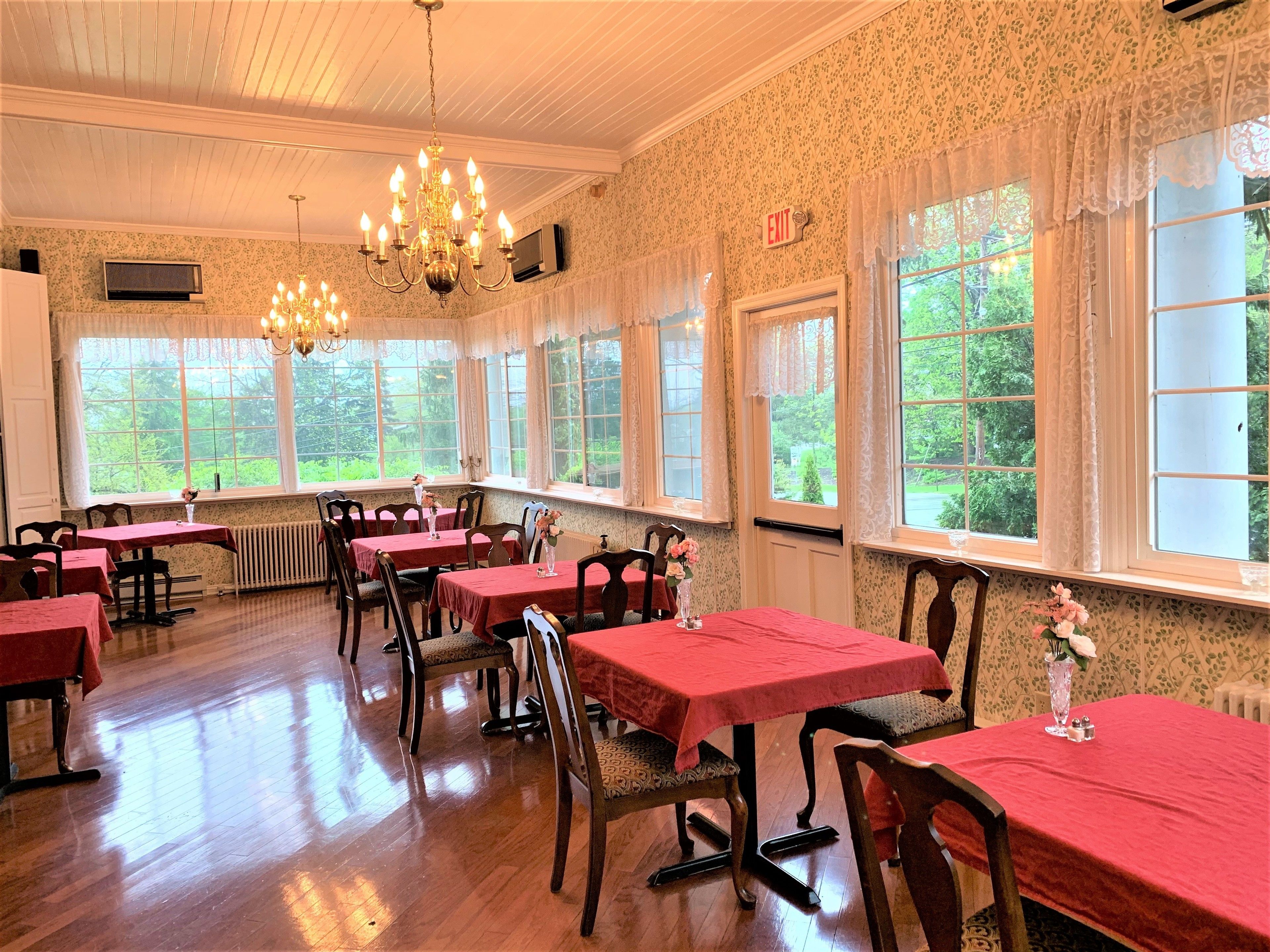 A dining room with multiple tables and chairs, large windows with curtains, and a chandelier.