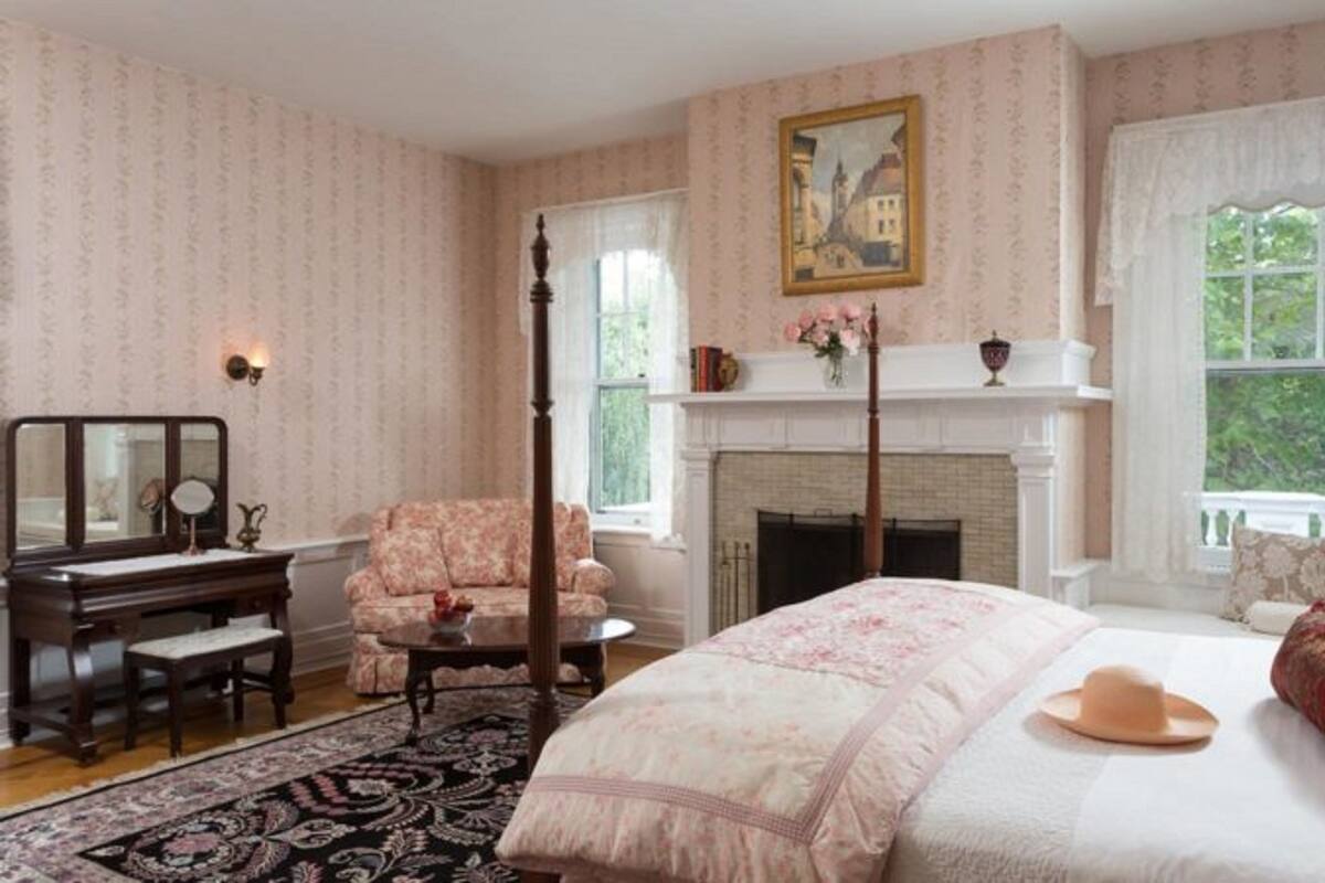 A bedroom with a four-poster bed, a fireplace, a desk with a mirror, and a chair.