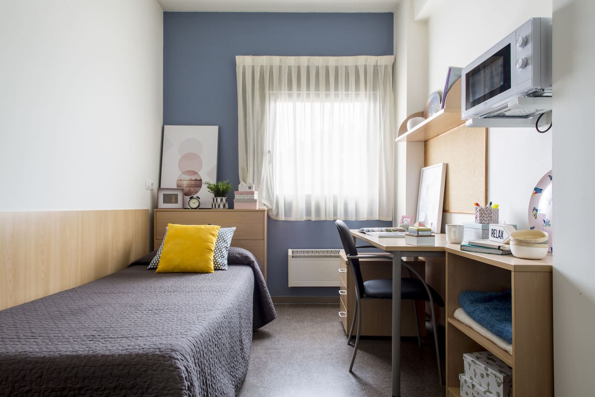Une chambre à coucher de taille réduite, comprenant un lit, un bureau, une chaise, un four à micro-ondes et une fenêtre avec des rideaux.