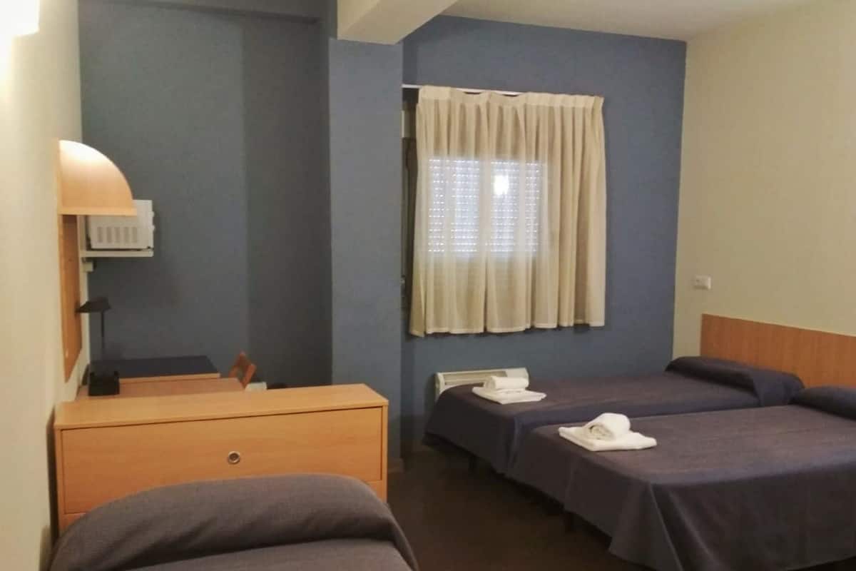 Une chambre d’hôtel avec deux lits simples, une commode en bois, un bureau avec une lampe et une fenêtre avec des rideaux.