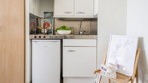 A compact kitchen with a refrigerator, stove, and a sink. There's a framed picture on the wall and a small table with a bowl of greens and a cup. - Residencia Universitaria Resa As Burgas - Centro Adscrito a REAJ - Campus Accomodation (Ourense)
