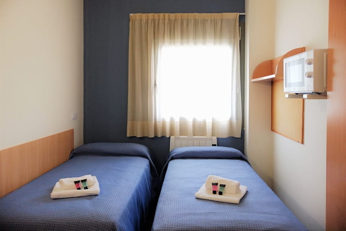 Une petite chambre d’hôtel avec deux lits simples, une fenêtre avec un rideau, un téléviseur fixé au mur et un radiateur.