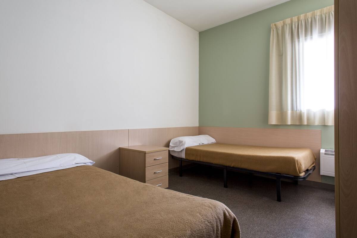 Une petite chambre propre, avec deux lits simples, une table de chevet en bois et une fenêtre avec des rideaux.