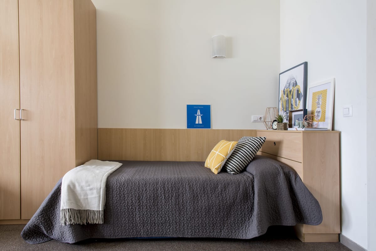 Un lit simple avec une couette grise, un plaid blanc et deux oreillers. Une petite table avec une lampe et des photos encadrées se trouve à côté du lit.