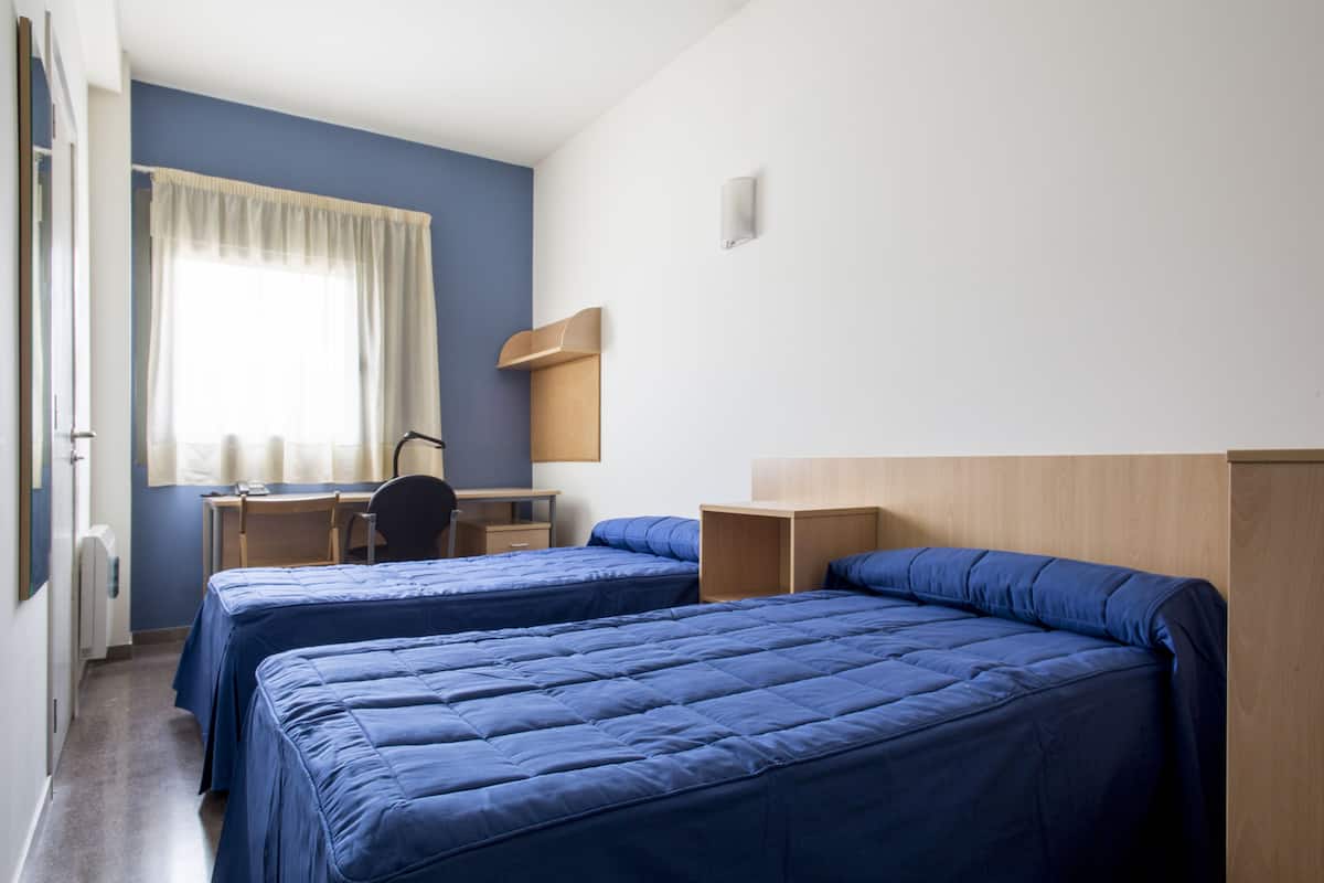 Une chambre d’hôtel avec deux lits, un bureau, une chaise et une fenêtre avec des rideaux.